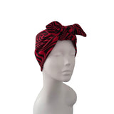 A red and black wavy patterned headwrap turban with a large bow, displayed on a white mannequin head against a plain white background.