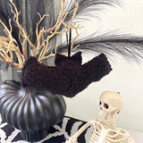 Decorative setup with black vase, black boucle fabric hanging bat, and skeleton figure on a striped surface.
