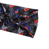 A close-up of a black fabric headband with a twisted knot, featuring a Halloween pattern of cobwebs, blue witches' hats, orange pumpkins, and small gold dots on a white background.