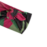 A close-up of a pleated fabric accessory, likely a headband or wrap, featuring a geometric pattern of magenta pink, olive green, and black stripes, tied in a knot on a plain white background.