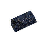 A dark navy blue fabric headband with a large twisted knot and an intricate floral pattern in muted gold and gray tones, shown on a plain white background.