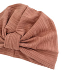 A close-up of a ribbed knit turban-style beanie in a dusty rose or mauve-brown color, shown on a plain white background.
