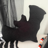 Black bat-shaped pillow on a white surface with a gray curtain in the background