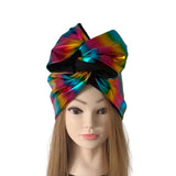A large metallic rainbow bow headband with black trim, displayed on a mannequin head with long light brown hair against a plain white background.