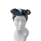 A dark teal fabric headband with a large bow, featuring a floral pattern in pink, orange, and white, displayed on a white mannequin head against a plain white background.