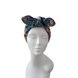 A dark teal blue fabric headwrap with a large bow, featuring a multicolor floral and leaf pattern in orange, pink, and yellow, displayed on a white mannequin head against a white background.