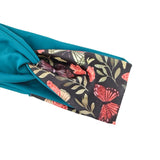 A folded teal fabric accessory, likely a scarf or bandana, showing both sides. One side is solid teal, while the other features a black background with a colorful butterfly and floral pattern in coral, green, and beige.