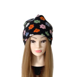 A black knit beanie hat with a colorful floral pattern, displayed on a mannequin head with long light brown hair against a plain white background.