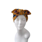 A mustard yellow fabric headwrap turban with a purple and blue floral pattern, displayed on a white mannequin head against a plain white background.