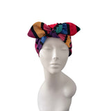 A multicolor patterned headwrap turban hat with a large bow, displayed on a white mannequin head against a plain white background.