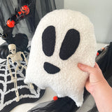 White ghost-shaped pillow with black eyes and mouth held by a hand, with Halloween decorations in the background.