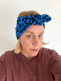 A woman wearing a bright blue and black leopard-print knotted headband, paired with a brown long-sleeve top, photographed against a plain white wall.
