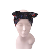 A black fabric headwrap or turban with a Halloween-themed print of orange pumpkins, spiders, and bats, styled on a light pink mannequin head against a white background.