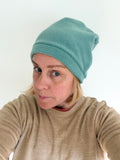 A woman wearing a soft, slouchy knit beanie in a muted teal-green color, paired with a beige crewneck sweater, photographed against a plain white background.