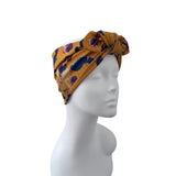 A mustard yellow fabric headwrap turban hat with a blue, purple, and orange abstract floral pattern, displayed on a white mannequin head against a white background.