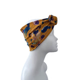 A mustard yellow knotted headband with a blue, purple, and orange abstract floral pattern, displayed on a white mannequin head in profile against a white background.