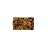 A mustard yellow ribbed fabric headband with a large twisted knot, decorated with a purple and teal floral pattern, shown on a plain white background.
