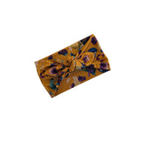 A mustard yellow fabric headband with a floral print in purple, blue, and green, tied in a knot at the center, shown on a plain white background.