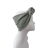 A sage green ribbed fabric headband with a large twisted knot detail, displayed on a white mannequin head in profile against a solid white background.