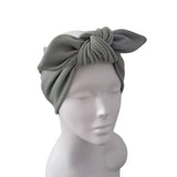 An olive green knitted turban-style headband with a large twisted bow, displayed on a white mannequin head against a plain white background.