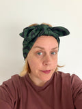 A woman wearing a dark green and black patterned knotted headband, paired with a dusty rose long-sleeve top, photographed against a plain off-white background.