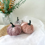 Pink velvet pumpkins with black stems on a white surface, with a vase of greenery in the background.
