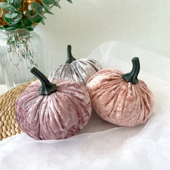 Two pink velvet pumpkins and a silver grey pumpkin on a white surface with a vase of greenery in the background.