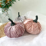 Two pink velvet pumpkins and a silver grey pumpkin on a white surface with a vase of greenery in the background.