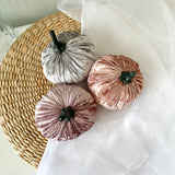 Three velvet pumpkins in gray, pink, and white on a woven mat.