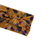 A mustard yellow ribbed fabric headband with a large twisted knot, featuring a purple and blue floral pattern, shown on a plain white background.
