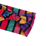 A colorful striped fabric headband with a large bow, featuring bold stripes of pink, teal, purple, yellow, and orange separated by black lines, on a plain white background.