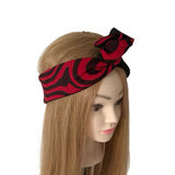 A red and black patterned fabric headband with a large bow, displayed on a mannequin head with long blonde hair against a plain white background.