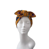 A mustard yellow fabric headwrap with a floral pattern in purple, blue, and green, styled on a white mannequin head against a plain white background.