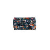 A fabric headband with a large front knot, featuring a detailed botanical and butterfly pattern in teal, pink, orange, and brown on a solid white background.