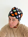 A woman wearing a black knit beanie with a colorful floral pattern, photographed in a casual selfie-style portrait against a plain light background.