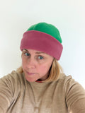 A woman wearing a two-tone knit beanie hat with a green top section and a dusty rose pink folded brim, paired with a beige knit sweater against a plain white background.