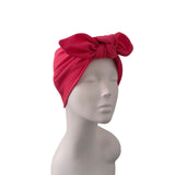 A bright red textured fabric headwrap turban hat with a large bow on top, displayed on a white mannequin head against a plain white background.