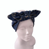 A navy blue fabric headwrap with a gold floral pattern, styled in a large bow on a white mannequin head against a plain white background.