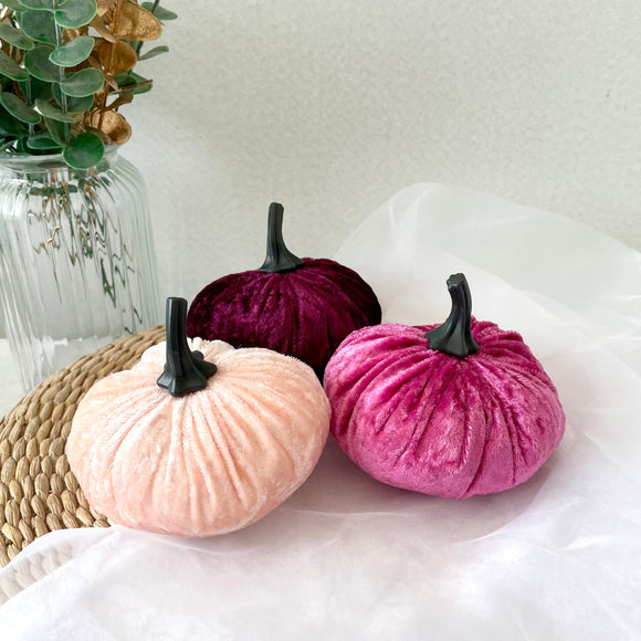 Three velvet pumpkins in pink, purple, and beige on a white surface with a vase of flowers in the background.