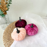 Three velvet pumpkins in pink, red, and beige on a white surface with a woven mat and plant in the background.