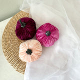 Three velvet pumpkins in purple, pink, and peach on a woven mat with sheer fabric.