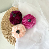 Three velvet pumpkins in purple, pink, and peach on a woven mat with sheer fabric.