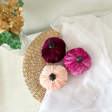 Three velvet pumpkins in purple, pink, and beige on a woven mat with sheer fabric and decorative elements in the background.