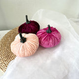 Three velvet pumpkins in pink, red, and beige on a white surface.