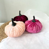 Three velvet pumpkins in pink, purple, and beige on a white surface.