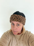A woman wearing a leopard print headband and a black beanie, taking a selfie against a plain white wall.
