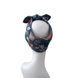 A dark teal bandana with a colorful floral and leaf pattern, styled on a white mannequin head against a plain white background.