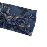 A close-up of a dark blue fabric headband with a distressed beige floral pattern and a knotted front detail, shown on a plain white background.