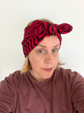 A woman wearing a red and black wavy patterned knotted headband, paired with a dusty rose long-sleeve top, photographed against a plain white background.